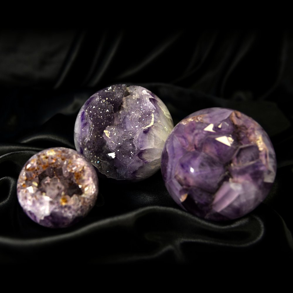 Three polished amethyst spheres with open crystal pockets, resting on black satin