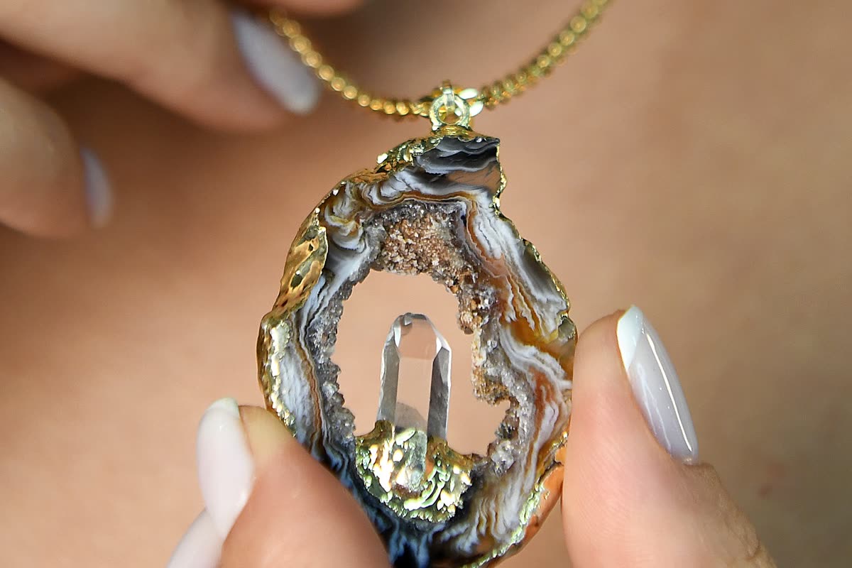 A banded agate slice pendant in a gold setting, with a clear quartz point mounted inside the open cavity, held between two fingers